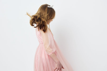 Playful young girl in a pink dress twirling, joyfully smiling, against a soft white background,...