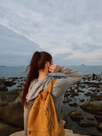 Young woman with a backpack enjoying a coastal view, wearing a gray hoodie, showcasing a sense of...