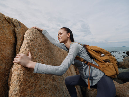 Young female hiker in sportswear climbing rocks, showcasing determination and adventure, with a backpack, against a cloudy backdrop, ideal for outdoor lifestyle conceptsの写真素材