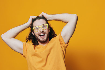 Happy young man with curly hair wearing yellow glasses and a bright orange shirt against a vibrant yellow background, expressing joy and excitementの写真素材