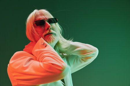 Stylish elderly man with long hair in sunglasses, posing against a gradient green background,...