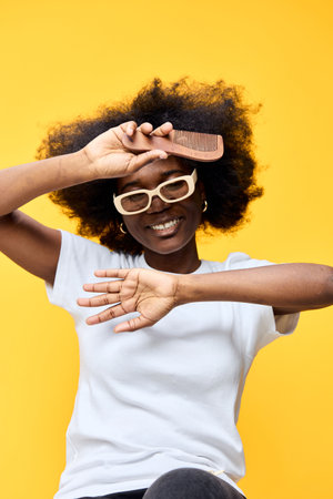 Happy young woman with natural afro hairstyle holding a comb, smiling playfully against a vibrant yellow background, embodying confidence and self careの写真素材