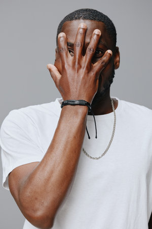 Portrait of a young man covering his face with his hand, wearing a minimalistic white t shirt, expressing emotion against a neutral gray backgroundの写真素材