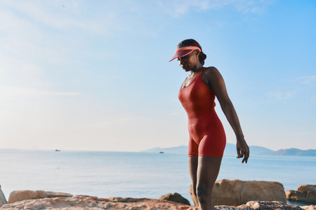 Stylish woman in a red swimsuit walking along the beach during sunset, with a serene ocean backdrop and gentle waves lapping at the shoreの写真素材