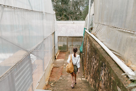 A young woman carrying a bag walks down the stairs in a greenhouse, showcasing sustainability,...