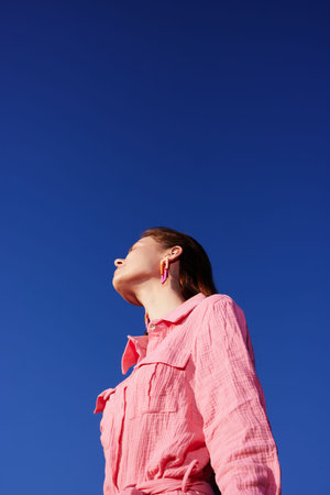 A young woman gazes upward against a clear blue sky, wearing a pink shirt that reflects a casual summer vibe and conveys a sense of freedom and aspirationの写真素材