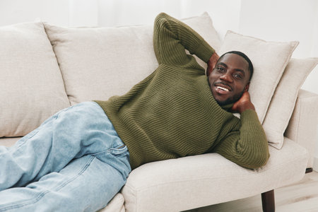 Cozy African man relaxing on a couch in a green sweater and jeans, expressing happiness with a...