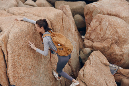 Young female climber in athletic wear navigating rocky terrain Her determination is evident as she climbs with a brown backpack on a sunny dayの写真素材