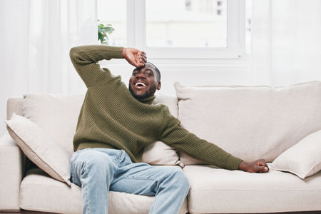 Happy young Black man in green sweater laughing on cozy sofa, enjoying his day at home with bright natural light and relaxed atmosphereの写真素材