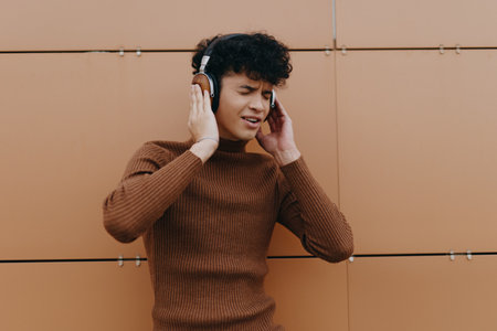 Fashionable young man listening to music on headphones in front of an orange wall with hands on headの写真素材