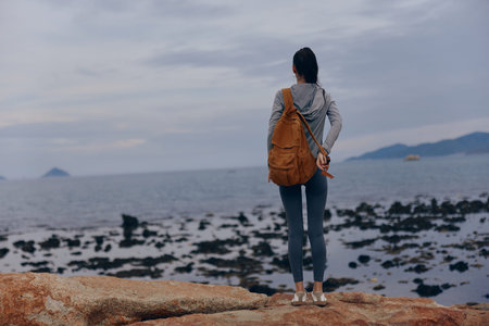 Young woman in casual grey hoodie and backpack gazing at the ocean, evoking a sense of adventure and tranquility in a coastal landscapeの写真素材