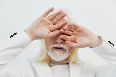 Portrait of a model with white hair and beard, covering face with hands in a minimalist white background, conveying a sense of mystery and calmnessの写真素材