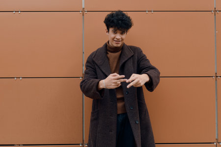 Fashionable young man in a brown coat with hands in his pockets, standing in front of an orange wallの写真素材