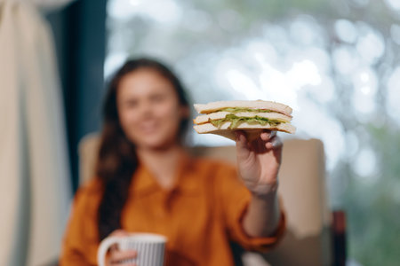 A woman holding a delicious sandwich with lettuce, sitting in a cozy chair, enjoying healthy eating in a bright, inviting setting, perfect for promoting healthy lifestylesの写真素材