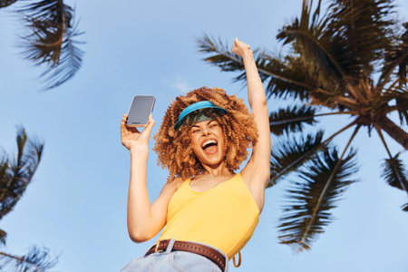 Cheerful woman celebrating outdoors under palm trees with a smartphone in hand Bright blue sky and...