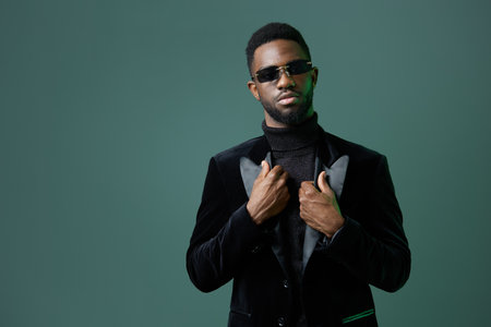 Confident man in sunglasses and black tuxedo jacket standing against vibrant green background,...
