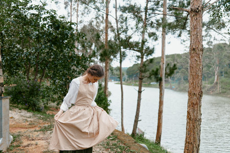 A young woman in a vintage dress stands by a serene lake, admiring her flowing skirt amid lush greenery, evoking a sense of tranquility and nature appreciationの写真素材