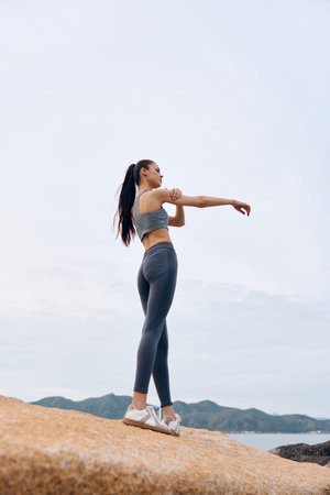 Fit young woman stretching outdoors in athletic wear by the sea at sunrise showcasing her healthy...