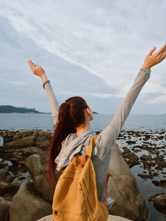 Young woman enjoying the ocean view with arms raised, wearing a casual outfit and a backpack, feeling free and connected to natureの写真素材