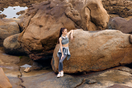 Young athletic woman in workout attire poses confidently on rocky coastline, showcasing strength and natural beauty in outdoor fitness settingの写真素材
