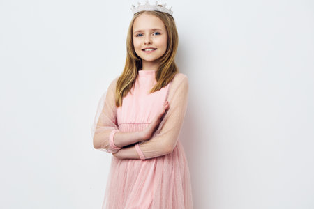 A cheerful young girl in a pink dress with tulle sleeves, wearing a tiara and smiling confidently against a simple white backgroundの写真素材