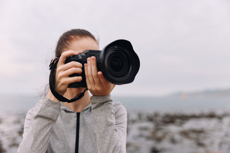 Young woman focused on photography near the beach, wearing a gray jacket, expression of concentration, capturing the beauty of nature, coastal sceneryの写真素材