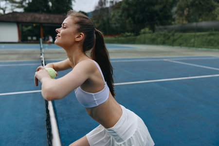 Young athletic woman in sportswear enjoying a moment after a tennis match, showcasing determination and joy on a sunny day on a blue tennis courtの写真素材