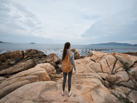 Woman with a backpack enjoying serene ocean view on rocky beach, wearing casual outfit, reflecting calm emotions in natural sceneryの写真素材