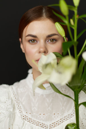 Elegant portrait of a young woman with green eyes holding white lilies against a dark background,...