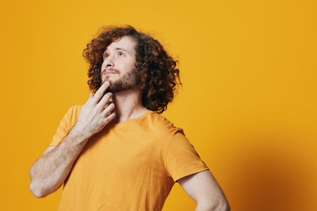 Thoughtful man with curly hair in an orange shirt, posed against a vibrant yellow background, contemplating ideas and showcasing a sense of curiosity and creativityの写真素材