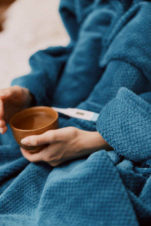 Person wrapped in blue blanket holding bowl with thermometer sitting on bedの写真素材