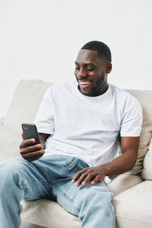 Smiling young Black man enjoying his time on a smartphone while sitting on a cozy couch, showcasing relaxed vibes and casual fashionの写真素材