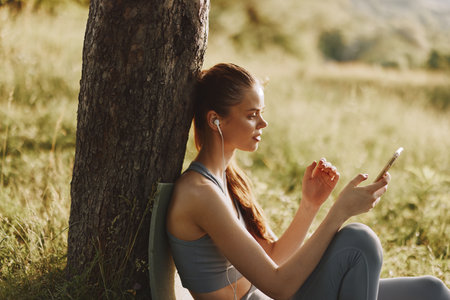 Young woman enjoying music outdoors in a fitness outfit, expressing joy and relaxation while using a smartphone near a tree in a sunny nature settingの写真素材