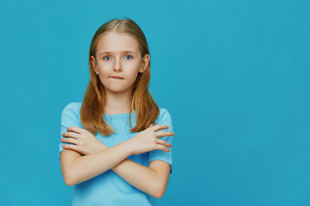 A young girl with long blonde hair wearing a light blue shirt, standing with crossed arms against a bright blue background, expressing a thoughtful emotionの写真素材
