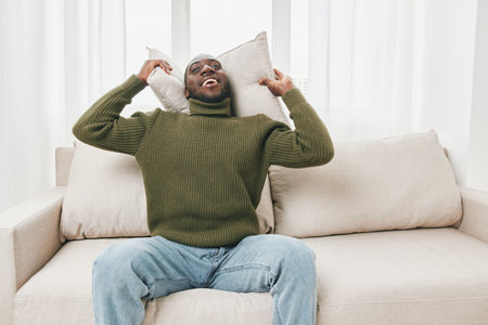 Happy Black man relaxing on a cozy sofa with pillows, wearing a green sweater, enjoying a peaceful moment indoors, conveying comfort and joyの写真素材
