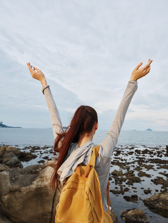 Young woman with long hair expressing joy against a scenic coastal background, celebrating freedom and nature, wearing casual attire with a backpackの写真素材