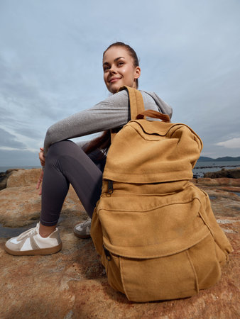Young woman with a backpack sitting on rocks by the sea, smiling in casual outdoor attire, capturing a sense of adventure and tranquilityの写真素材