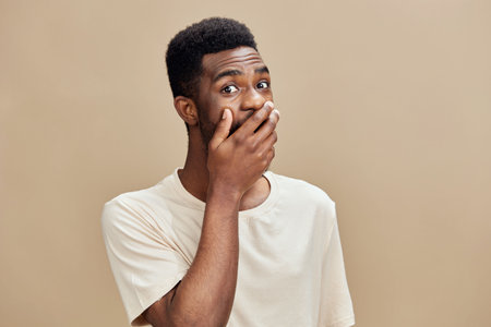 Surprised Black man covering his mouth with his hand against a neutral beige background, expressing shock or disbelief, wearing a casual light colored t shirtの写真素材