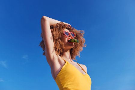 A woman with curly hair wearing heart shaped sunglasses holds a cucumber in her mouth against a bright blue skyの写真素材