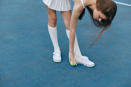 Young female tennis player in a white sports outfit picking up a tennis ball on a blue court, showcasing athleticism and focus in her expressionの写真素材
