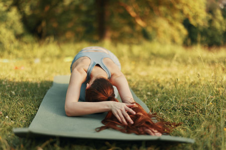 Young woman practicing yoga outdoors, wearing grey athletic wear, in a peaceful pose on a green mat surrounded by greenery, evoking tranquility and connection with natureの写真素材