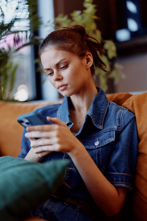 Young woman in denim shirt sitting on couch looking at phone, casual lifestyle conceptの写真素材