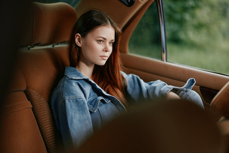 A young, stylish woman with long brown hair wearing a denim jacket, seated in a vintage car, exuding...