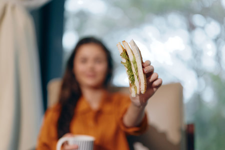 Young woman holding a sandwich in a cozy interior, conveying a healthy lifestyle and enjoyment of fresh food with a blurred backgroundの写真素材