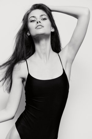 Elegant young woman in black swimsuit posing confidently against a soft gray background, showcasing beauty and grace with flowing hair. Fashion and modeling concept.の写真素材