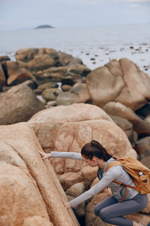 Young female climber overcoming rocky terrain by the seaside, showcasing determination and outdoor adventure in a gray athletic outfitの写真素材