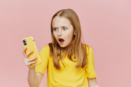 Surprised girl in yellow t shirt looking at smartphone against pink background, expressing shock and excitementの写真素材