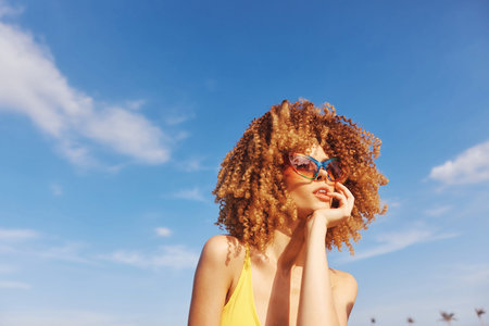Young woman with curly hair in sunglasses enjoying sunny weather against a blue skyの写真素材