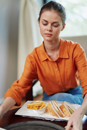 Young woman in an orange shirt preparing a healthy meal with sandwiches and fries, emphasizing fresh ingredients and vibrant colors in a cozy environmentの写真素材