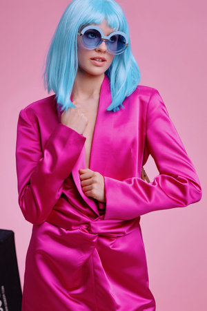 Bright pink suit, blue hair, and blue sunglasses, woman on pink background holding a black bagの写真素材